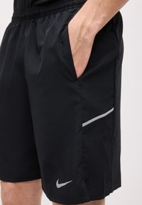 Άνδρας που φοράει μαύρα αθλητικά σορτς Nike με το χέρι στην πλαϊνή τσέπη και ανακλαστικές λεπτομέρειες κοντά στο τελείωμα.