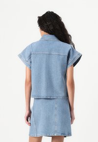 Jachetă din denim de culoare albastru deschis, cu mâneci scurte, prezentată împreună cu o fustă din denim asortată. Linii curate și detalii de cusătură vizibile.
