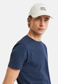 Cappello bianco con logo ricamato "GANT", indossato con una camicia a maniche corte blu navy. Il cappello ha un design strutturato e una visiera curva.
