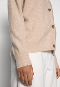 Vaaleanbeige neulottu neuletakki, jossa on resorit hihansuissa, viisi puisesta napista ja pehmeä pinta. Päällä vaaleat housut.