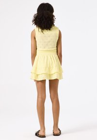 Robe sans manches jaune avec un design en œillets floraux, taille élastique et jupe à volants superposés. Le mannequin porte des sandales noires, les cheveux en boucles naturelles.
