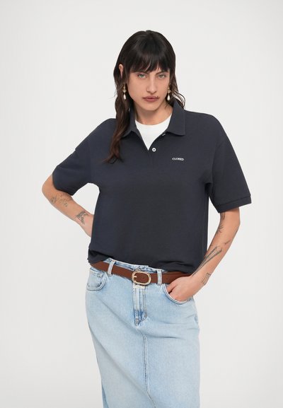 Γυναίκα με σκούρα μαλλιά φορώντας μπλε κοντομάνικο πόλο μπλουζάκι, ανοιχτόχρωμη τζιν φούστα, καφέ ζώνη και χρυσά σκουλαρίκια, με τατουάζ και στα δύο χέρια.