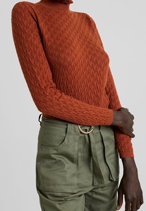 Femme portant un pull à col roulé en maille torsadée couleur rouille et un pantalon taille haute vert olive avec une ceinture en tissu et une boucle en anneau doré.