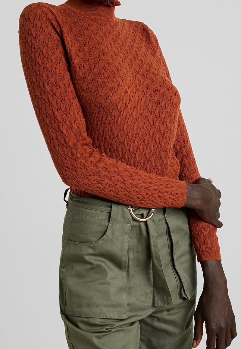 Femme portant un pull à col roulé en maille torsadée couleur rouille et un pantalon taille haute vert olive avec une ceinture en tissu et une boucle en anneau doré.