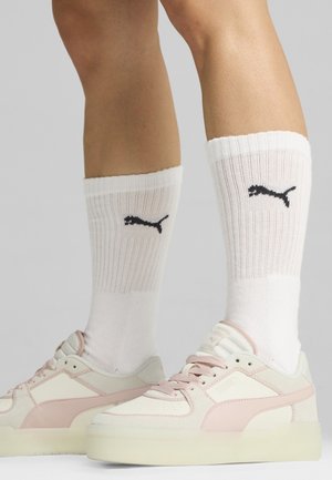 Beine, die weiße Puma Crew-Socken und cremefarbene sowie rosa Puma-Sneaker tragen, stehen auf einer hellgrauen Oberfläche.