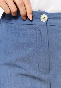 Gai zilie džinsi ar pogu aizdari, gludu tekstūru un priekšējo kabatu ar maziem šuvju rakstiem.