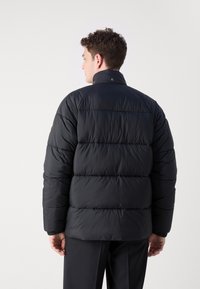 Zwarte pufferjas met een hoge kraag, horizontale naden, elastische manchetten en een gladde textuur. Beschikt over drukknoopsluitingen op de kraag.