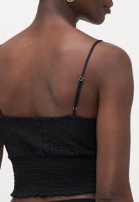 Svart croppet topp med tynne justerbare stropper, teksturert hekledesign, strikket midje og volanger på kanten, som fremhever intrikate mønstre.