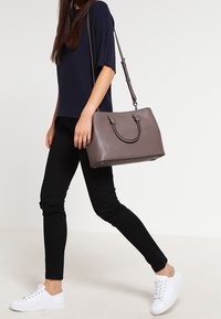 Grijze leren handtas met gestructureerd ontwerp, dubbele handvatten en een afneembare schouderriem. Outfit bestaat uit een marineblauw shirt, zwarte broek en witte sneakers.