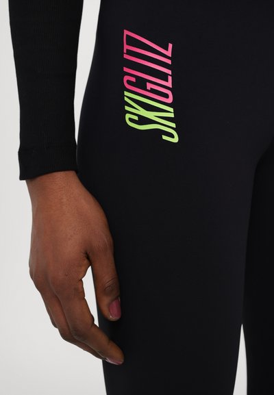 Fekete leggings rugalmas anyagból, színes, átlós pink és zöld logóval a combon, mellette egy kéz közeli felvétele.