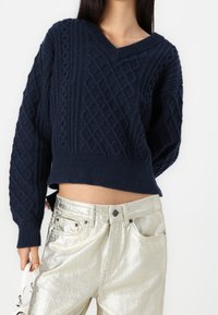 Granatowy sweter dziany z dekoltem w serek, z fakturowanymi wzorami w warkocze i ściągaczami u mankietów, połączony z metalicznie srebrnymi spodniami o wysokim stanie.
