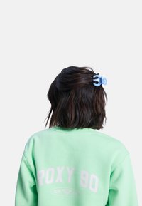 Una pinza de cabello azul claro asegurando el cabello oscuro, que se lleva con un sudadera verde menta que tiene texto en blanco en la parte posterior. Textura suave y diseño casual.