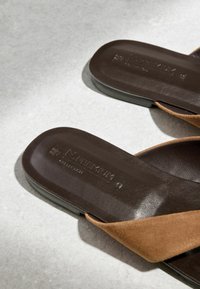 Sandales slide en cuir marron avec larges brides en daim sur une surface gris clair, montrant un gros plan du détail de la semelle intérieure et de l'embossage de la marque.