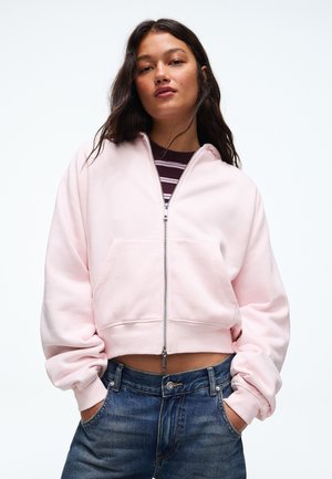 Une femme porte un sweat à capuche rose pâle zippé par-dessus une chemise rayée et un jean bleu. Elle se tient debout, les mains dans les poches, sur un fond clair.