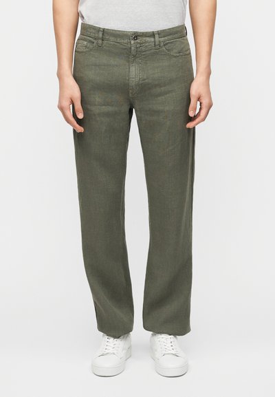 Homme portant un pantalon droit vert olive, des baskets blanches et un t-shirt gris clair rentré, debout avec les mains détendues le long du corps.