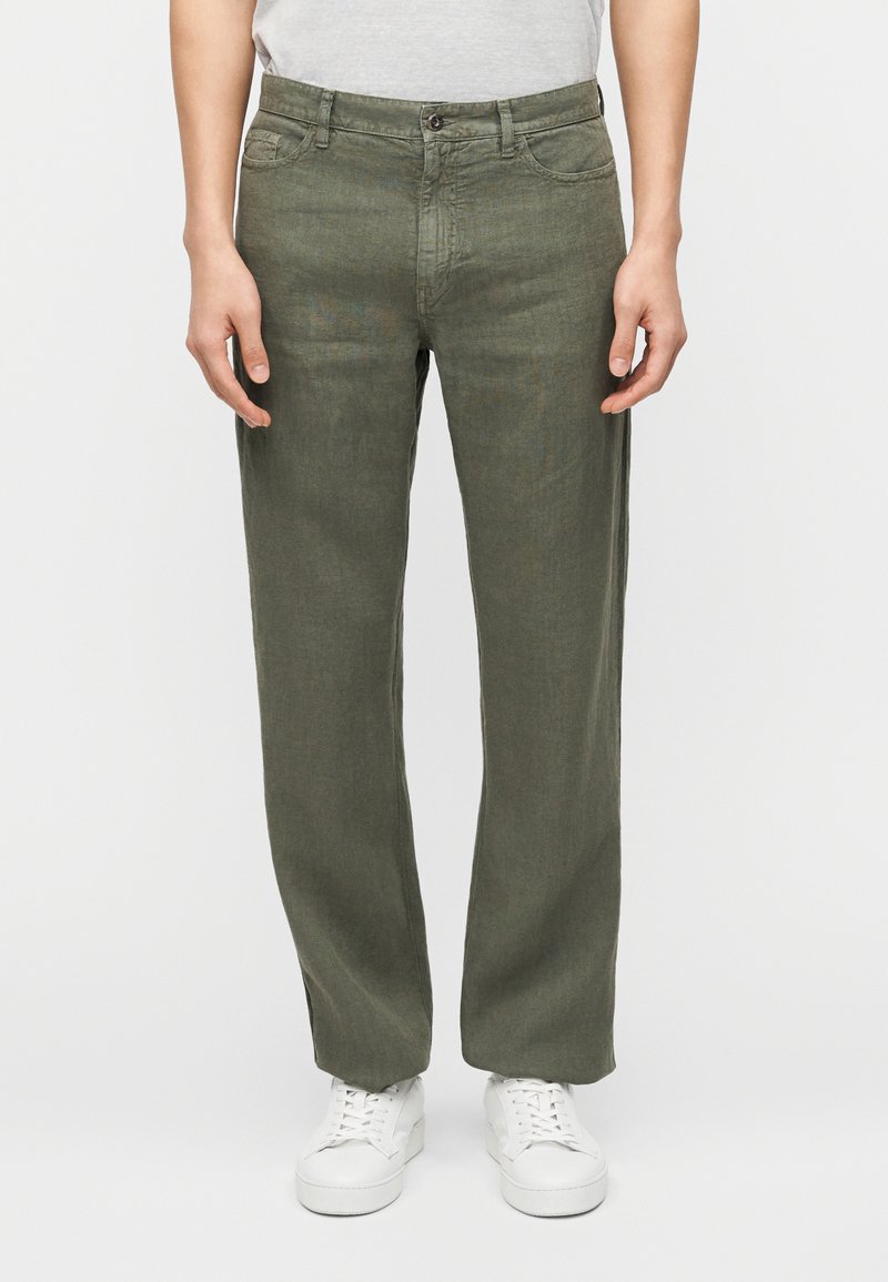 Άνδρας που φοράει ελιάς πράσινο πα ντελόνι απευθείας γραμμής, λευκά αθλητικά παπούτσια και ανοιχτογκρι μπλουζάκι με γιακά tucked μέσα, στέκεται με χαλαρά τα χέρια στα πλευρά.