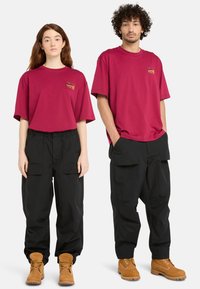 Camisetas de gran tamaño en color burdeos con un pequeño logo, combinadas con pantalones cargo negros holgados y botas marrones con cordones. Material de algodón texturizado.