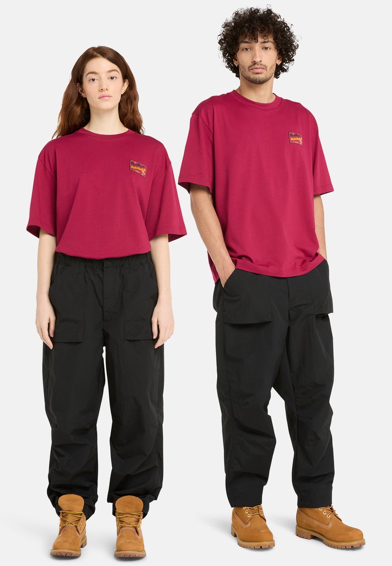 Camisetas de gran tamaño en color burdeos con un pequeño logo, combinadas con pantalones cargo negros holgados y botas marrones con cordones. Material de algodón texturizado.