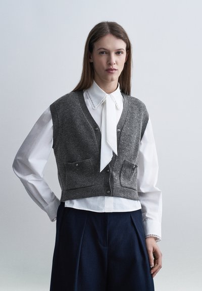 Jeune femme portant un chemisier blanc avec col en dentelle et ruban, un gilet en tricot gris boutonné avec des poches, et un pantalon large sombre, debout devant un fond uni.