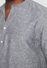 Harmaa pellavapaita, jossa on mandariinikaulus, kolme nappia kauluksella ja hienovarainen tekstuuri kankaassa. Saumoissa näkyvät ompeleet.