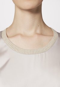 Light-colored blouse with a smooth texture and a ribbed, beige collar. The neckline is round and sits close to the neck.