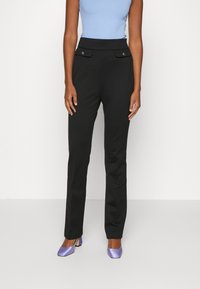 Černé kalhoty na míru s hladkou texturou, vybavené předními kapsami s ozdobnými knoflíky, doplněné světle modrým topem a fialovými podpatky.