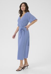 Vestido envolvente azul claro con mangas cortas, cintura ceñida con cinturón y un diseño fluido hasta el tobillo. Combinado con sandalias de tacón negro con tiras.