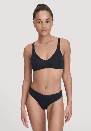 Woman with hair in bun wearing black bralette and black underwear with lace trim, standing against plain light background.