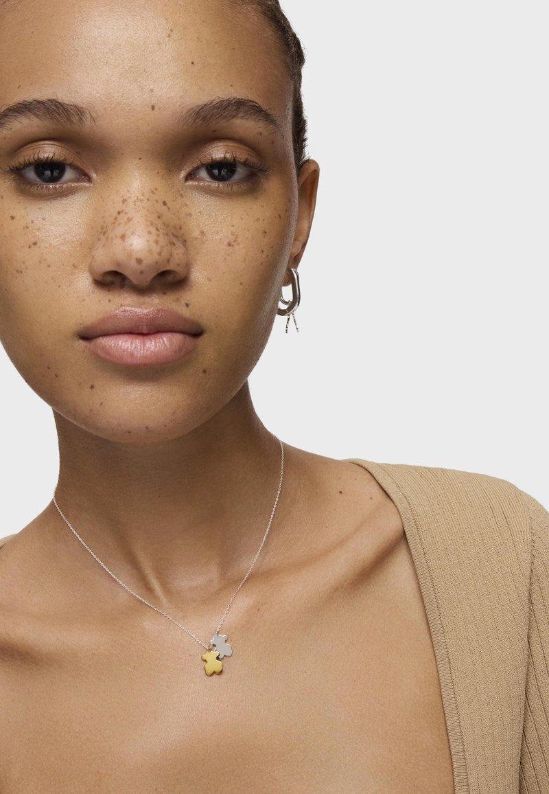 Collana d'argento con due pendenti a forma di stella, uno dorato e uno argento, indossata su un collo nudo. Il modello ha la pelle di colore marrone chiaro e lentiggini.