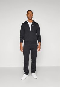 Melns rāvējslēdzēja sporta džemperis un atbilstoši sporta bikses no mīksta auduma, ar sānu kabatām, ribveida aprocēm un logotipu krūtīs, nēsāts ar baltām kurpēm.