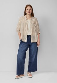 Beżowa i kremowa w paski koszula z krótkim rękawem, zapinana z przodu, zestawiona z szerokimi granatowymi dżinsami i białymi klapkami.