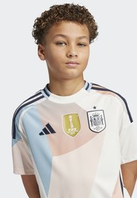 Jeugdvoetbalshirt in het wit met pastel geometrische patronen. Bevat een FIFA 2023 Champions badge en een embleem van het Spaanse nationale team. Zwarte accenten.