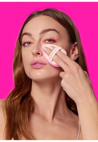 Make-up spons met een zachte, ronde vorm en een roze accent. Het model brengt product aan op haar wang tegen een felroze achtergrond.