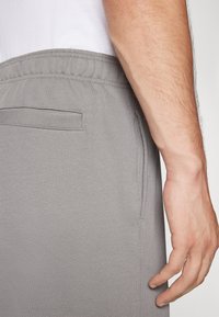 Pantalones jogger grises con cintura elástica, tejido texturizado y un bolsillo lateral, mostrados junto a una mano descansando sobre el material.