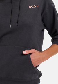 Schwarzer Hoodie mit einer vorderen Tasche, Kordelzugkapuze und kleinem ROXY-Logo in verlaufendem Pink auf der Brust. Weicher Stoff.