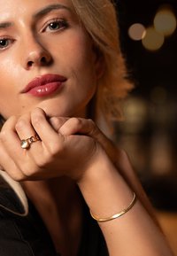 Kultainen rannekoru ja sormus, joissa on pyöreä kivi. Lähikuva kasvojaan koskettavista käsistä, jotka ovat sileitä ja joissa on vaaleat hiukset, neutraali tausta.