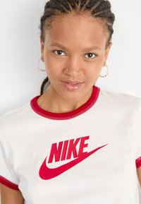 Valkoinen t-paita, jossa on punaiset ringer-kaulus ja -hihansuut, etuosassa on suuri punainen Nike-logo, valmistettu pehmeästä ja sileästä kankaasta.