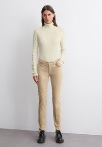 Beige housut, joissa on keskikorkea vyötärö, yhdistettynä vaalean kermaiseen ribattuhihoiseen turtleneck-paitaan. Asusteena mustat nauhakenkät.