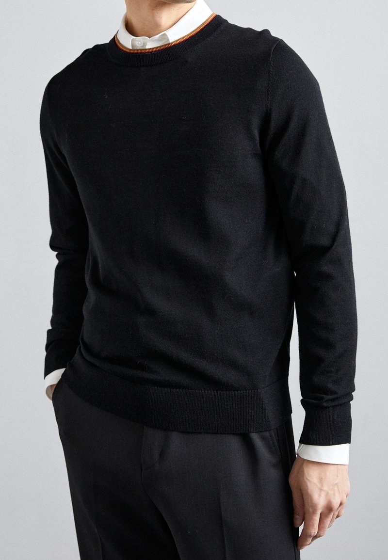 Homme portant un pull noir à col rond par-dessus une chemise blanche à col avec un liseré marron, assorti à un pantalon noir, mains dans les poches.