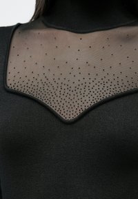 Haut noir à col montant avec un panneau en mesh transparent orné de perles noires éparses, créant un design texturé au niveau de la poitrine.