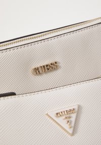 Weißer strukturierten Handtasche mit goldenen Beschlägen und geprägtem "GUESS"-Logo. Enthält ein dreieckiges Logo-Patch mit einem goldenen Akzent.