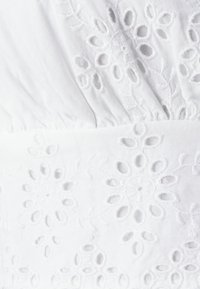 White fabric featuring intricate floral embroidery with cut-out patterns, showing a textured appearance and gathered detailing at the waist.