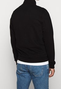 Svart sweatshirt med hög krage, tillverkad av mjukt tyg. Har ribbat avslut och muddar, bärs över en vit skjorta, kombinerad med blå jeans.