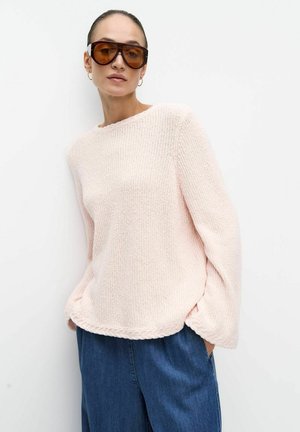 Kvinde iført store ravfarvede solbriller, lys rosa striktrøje med tekstureret kant og løse blå cowboybukser, stående mod en hvid væg.