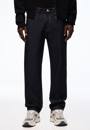 Person trägt schwarze Jeans mit geradem Bein, weiße und graue Sneakers und eine schwarze Jacke mit Reißverschluss, steht mit den Händen an den Seiten.