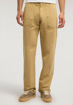 Uomo che indossa pantaloni beige a gamba dritta con grandi tasche frontali e sneakers casual marroni, in piedi davanti a uno sfondo semplice.