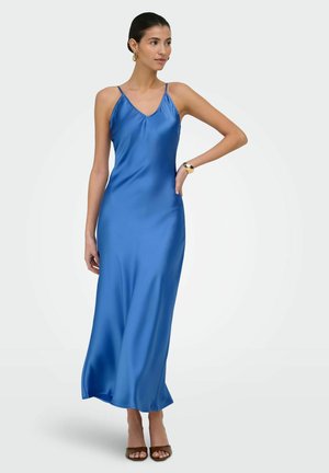 Nő áll egy kézzel a csípőjén, kék ujjatlan szatén maxiruha és barna nyitott orrú magas sarkú cipő van rajta, egyszínű háttér előtt.