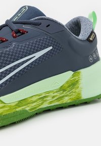 Gros plan d'une chaussure de trail bleu marine et verte avec une semelle intermédiaire texturée, des lacets rouges et une étiquette Gore-Tex sur le col rembourré.