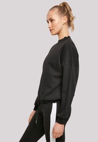 Schwarzer Pullover mit lässigem Schnitt und gerippten Bündchen, mit einem Kordelzugsaum. Kombiniert mit schwarzen Hosen, akzentuiert durch weiße Streifen.