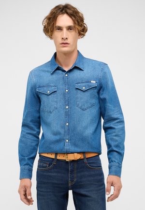 Blue denim shirt with button-down collar, two chest pockets, and white buttons. Worn with dark jeans and a tan belt. Smooth texture.